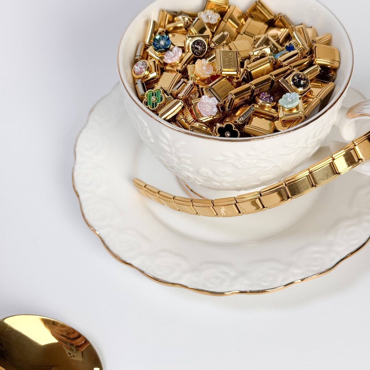 White teacup filled with gold jewelry pieces on a white saucer with a gold spoon.